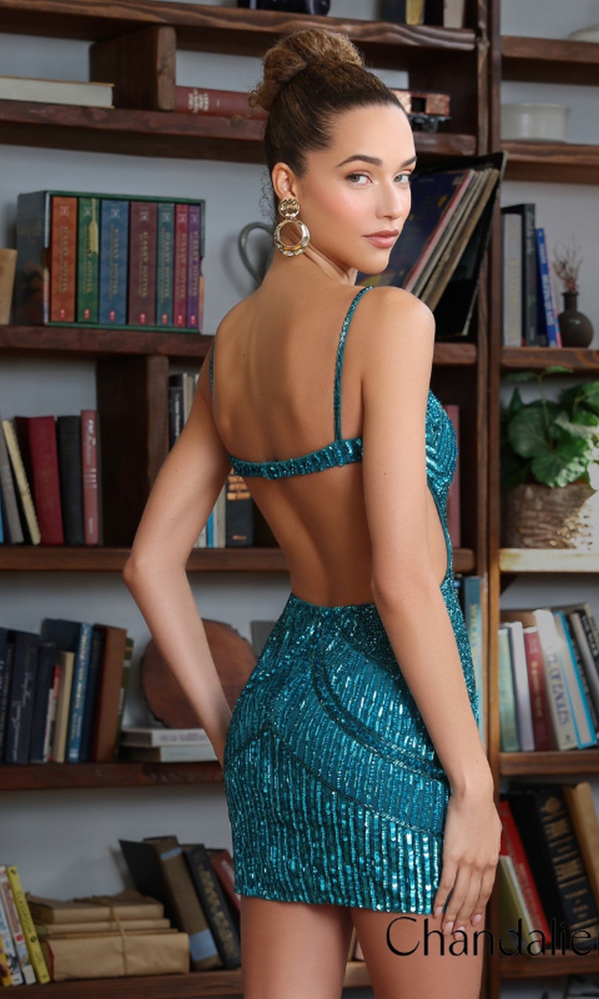 Woman stands in front of bookshelves wearing the Chandalier 770092 Short Prom Dress, a sparkly blue sequin dress with an open back. Her hair is styled in a bun, she wears large hoop earrings, and looks over her shoulder at the camera.